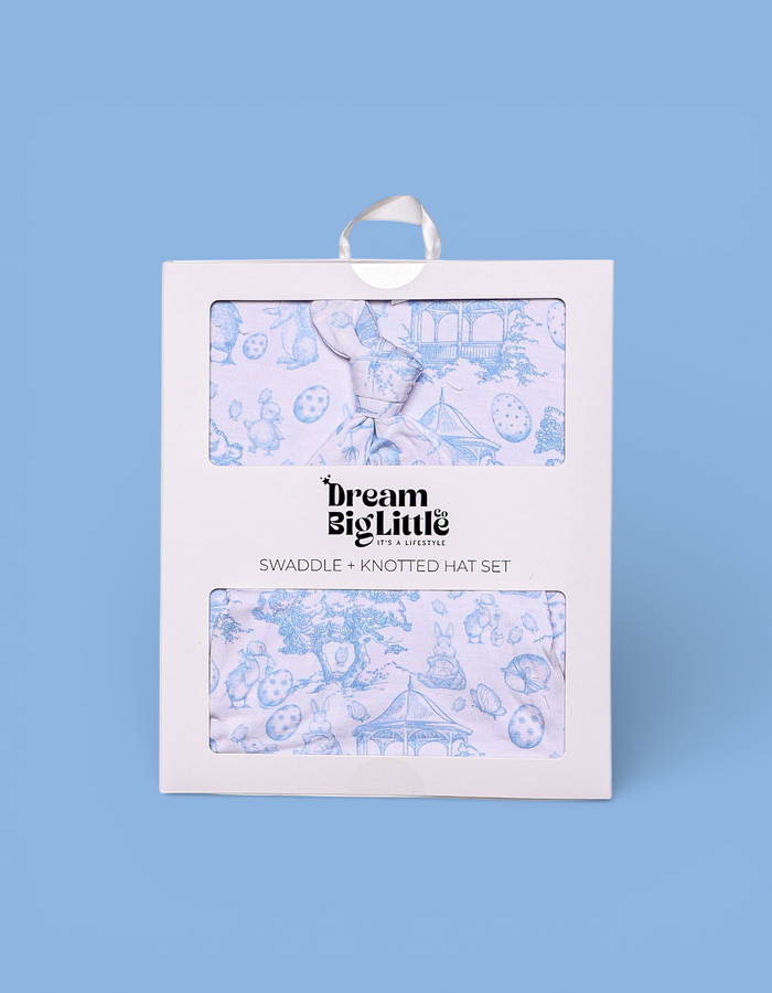 Packaged swaddle and knotted hat set with blue toile pattern on pale pink fabric, displayed in white retail box.