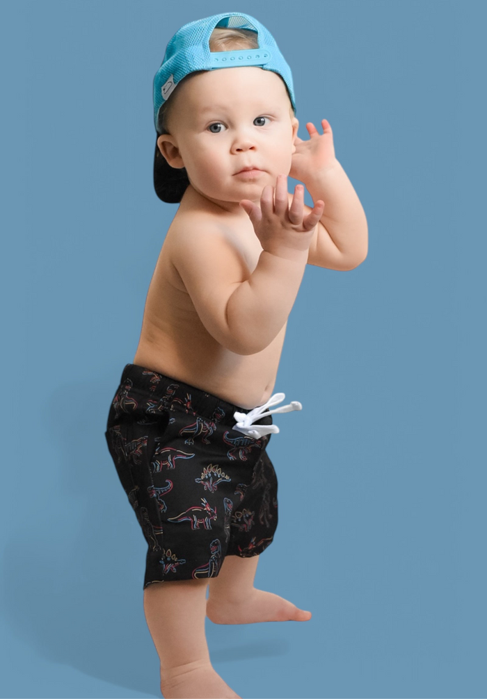 Toddler wearing black swim trunks with colorful dinosaur and plant print on light blue background.