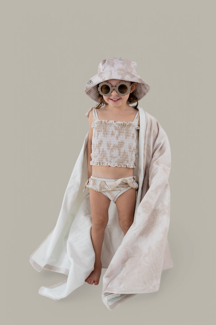 Child wearing cream-colored towel draped around shoulders over matching swimwear and bucket hat.