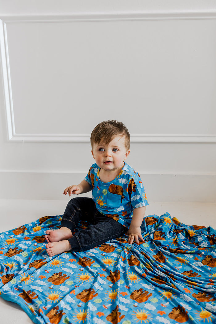 Toddler wearing blue short-sleeve tee with bear and sky print, sitting on matching patterned fabric.