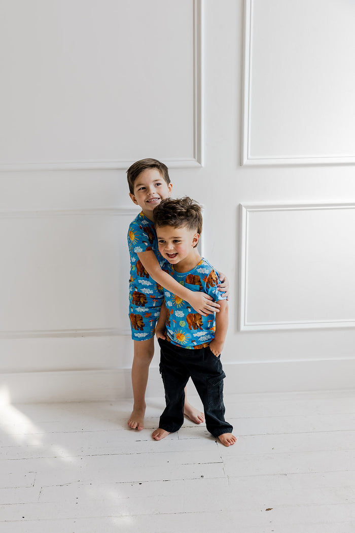 Two young boys wearing blue short-sleeve t-shirts with bear and cloud print, paired with dark pants.