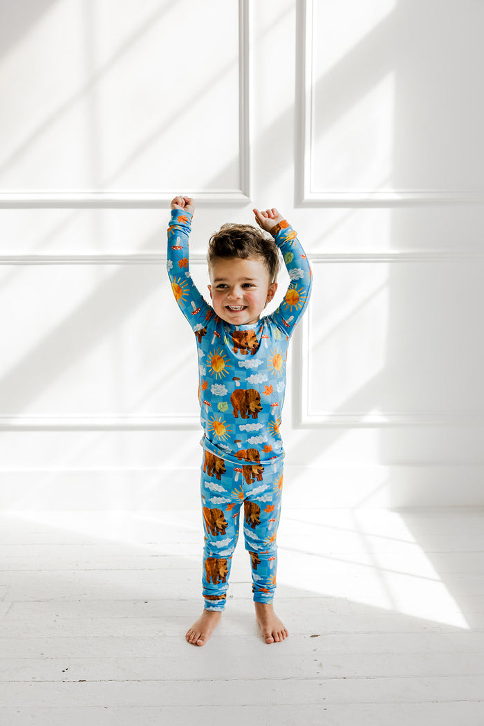 Child wearing blue pajama set with bear, sun, and cloud print throughout long sleeves and pants.
