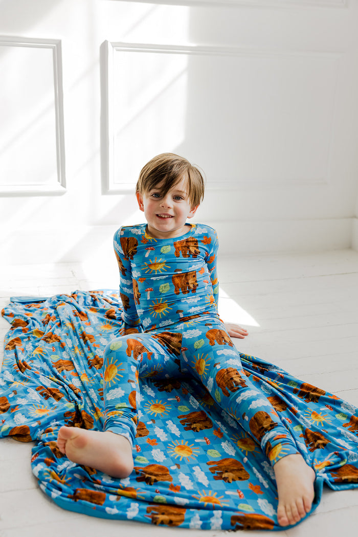 Toddler wearing blue pajamas with brown bear and sun print, sitting on matching bedding set.