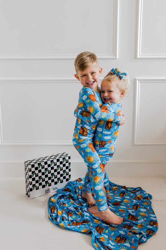 Two children wearing blue pajamas with bear and sun print, one child holding a coordinating blanket.