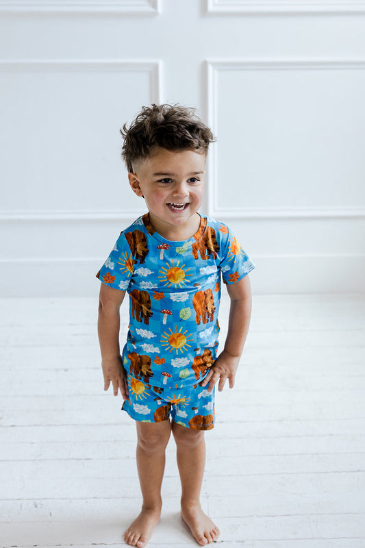 Toddler wearing blue short-sleeve shirt and shorts with bear, sun, and cloud print.