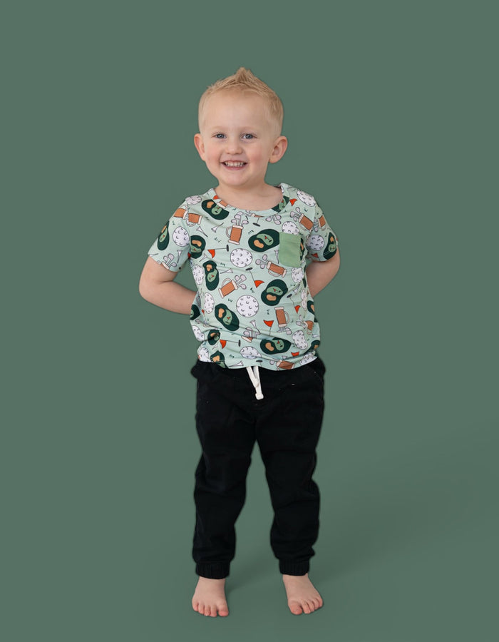 Toddler wearing cream short-sleeve tee with golf-themed print and green chest pocket, paired with black joggers.