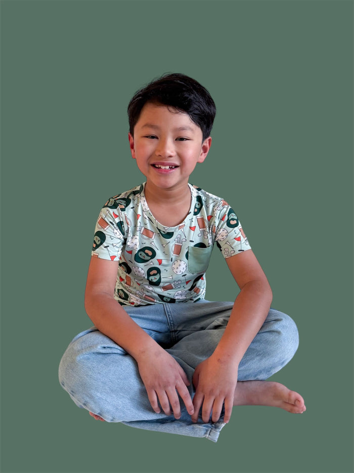 Child wearing short-sleeve pocket tee with golf-themed print in cream, navy, and red tones, paired with light blue jeans.