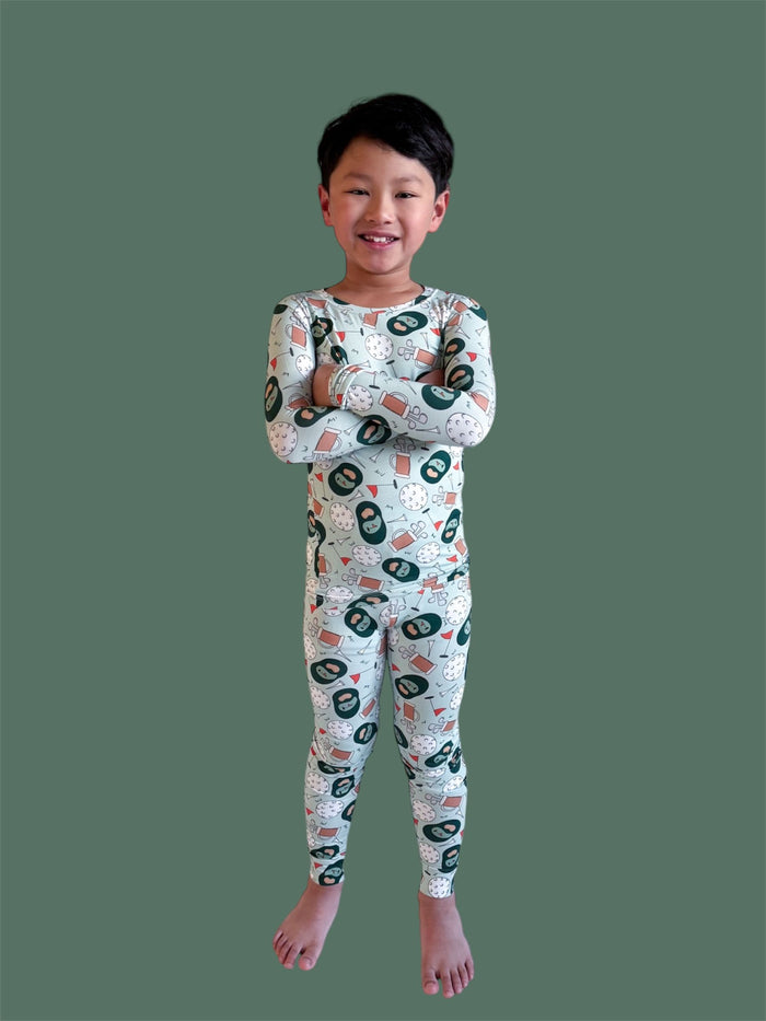 Child wearing long-sleeved pajama set with golf-themed print including clubs, balls, and flags on cream background.