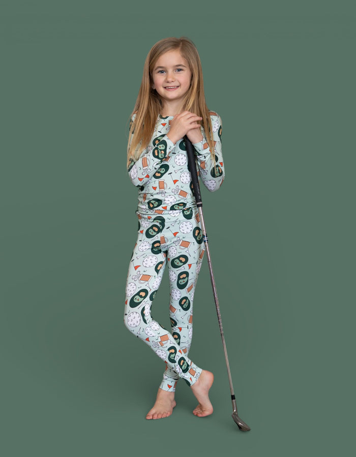 Child wearing white long-sleeve top and leggings with golf-themed print, holding a golf club against green background.