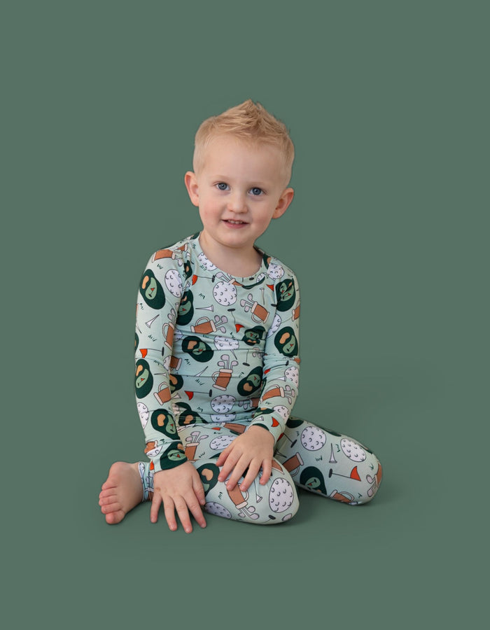 Toddler wearing long-sleeve pajamas with golf-themed print including clubs, balls, and flags on sage green background.