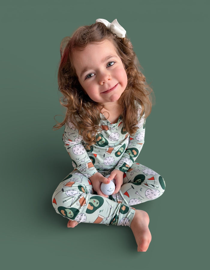 Young child wearing cream pajamas with golf-themed print including golf balls, clubs, and flags, with white bow hair clip.