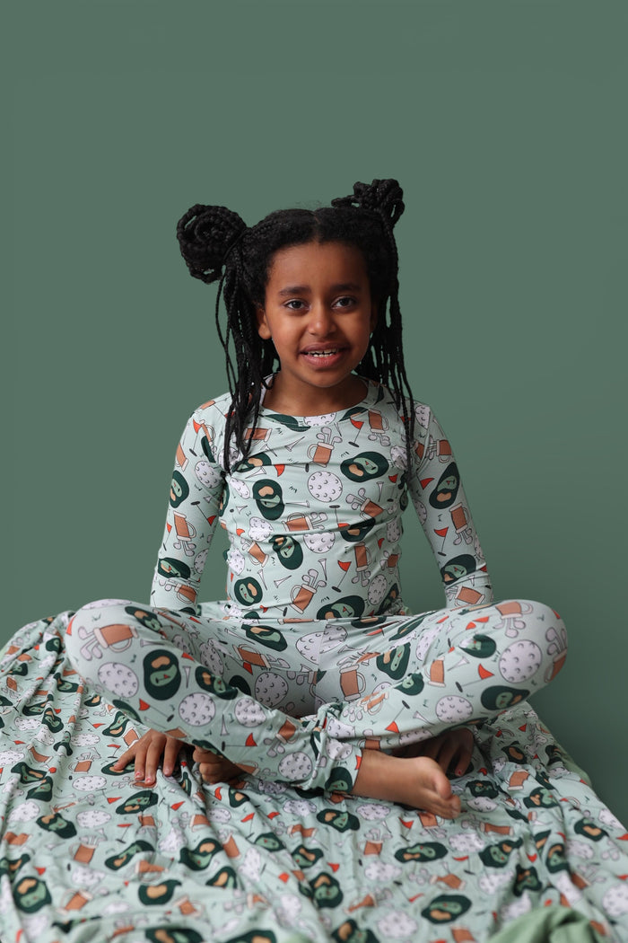 Child wearing long-sleeve pajama set with golf-themed print including clubs, balls, and flags on sage green background.