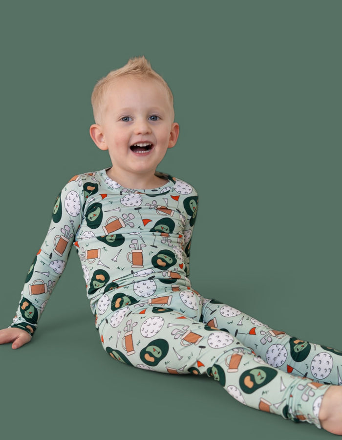 Toddler wearing long-sleeve pajamas with golf-themed print including golf balls, clubs, and flags on sage green background.