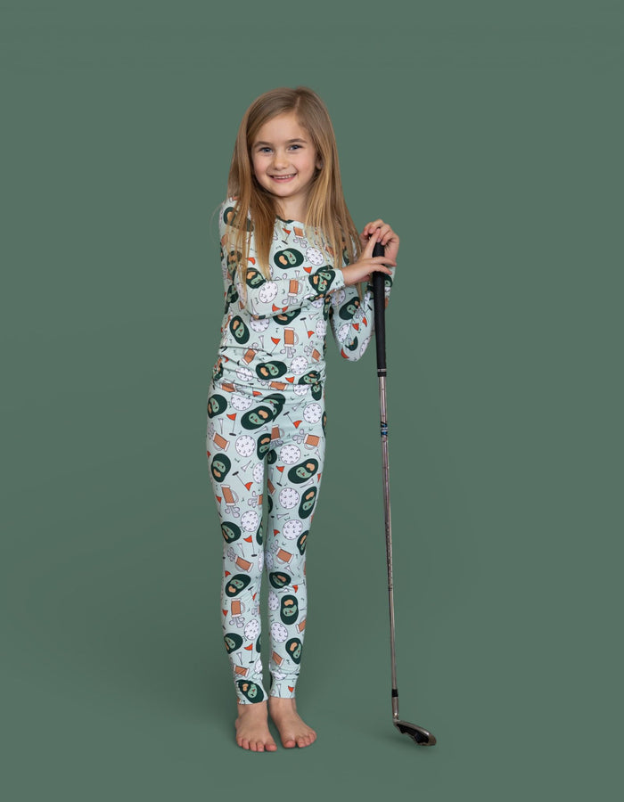 Child wearing white two-piece pajama set with golf-themed print, holding a golf club.
