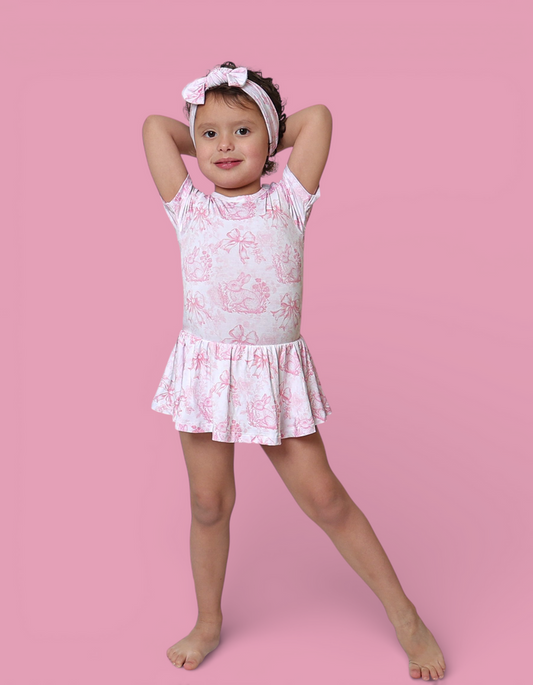 Child wearing white sleeveless bodysuit dress with pink toile pattern, matching headbow, on pink background.