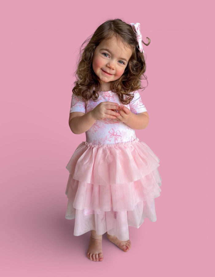 Toddler wearing pink short-sleeved top with toile print and layered pink tulle skirt with bow detail.