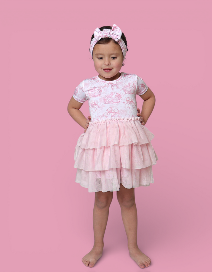Child wearing pink and white toile print short-sleeve top with tiered tulle skirt and matching bow headband.