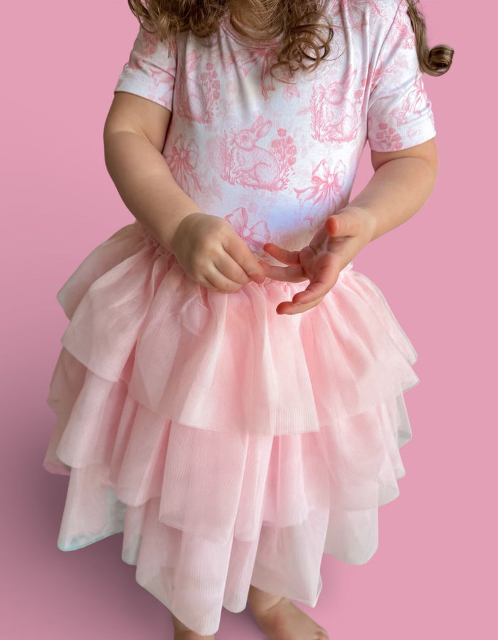 Toddler wearing pink and white toile print short-sleeve top with layered pink tulle skirt against pink background.