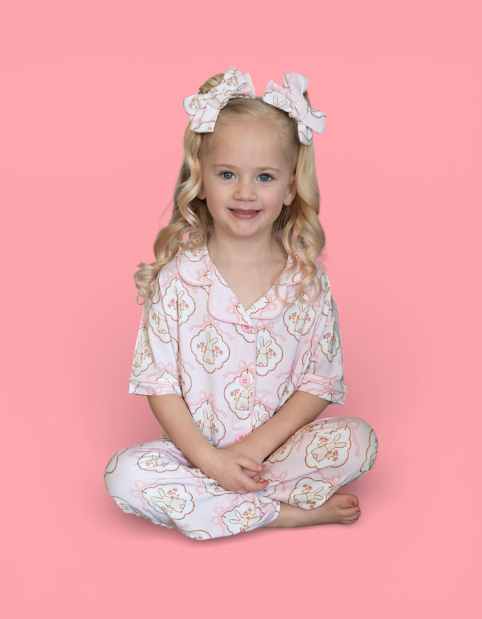Young girl wearing white pajama set with bunny pattern, matching hair bows, seated against pink background.