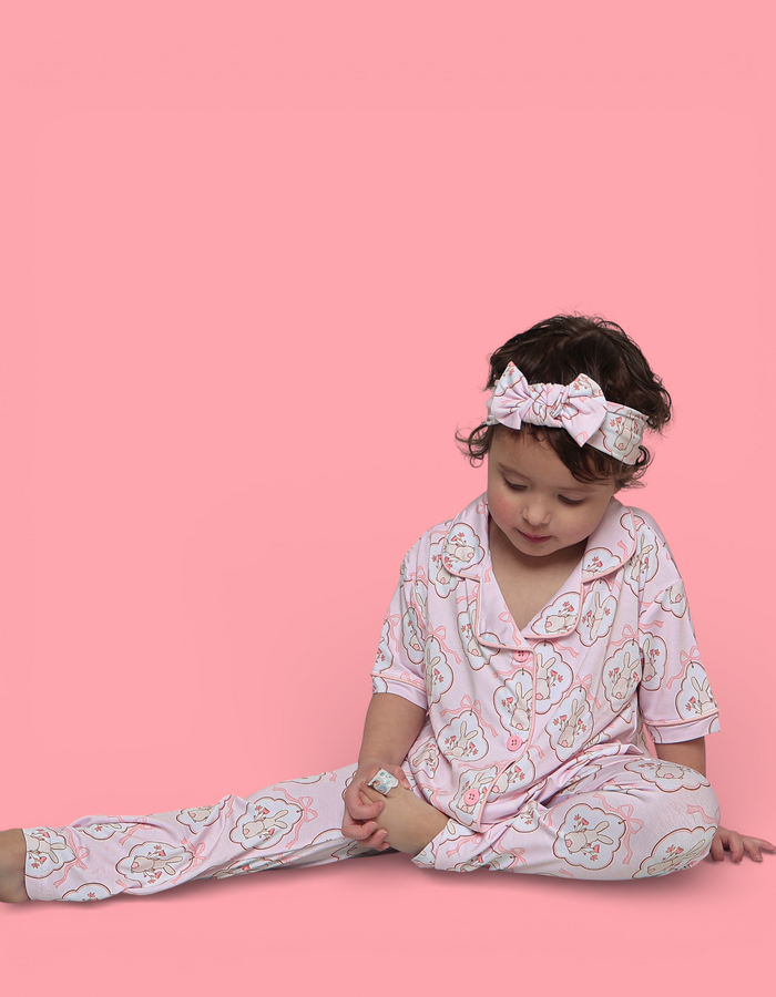 Young child wearing pink pajama set with bunny print and matching bow headband against pink background.