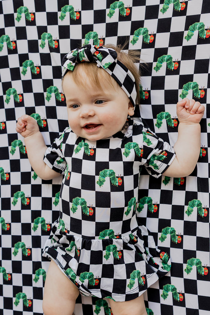 Infant wearing black and white checkered bodysuit dress with green caterpillar and red apple print, matching headband.
