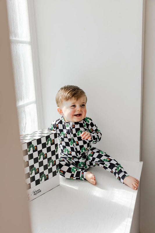 Toddler wearing black and white checkered romper with green caterpillar print, sitting by a window.