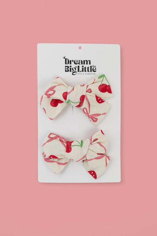 Two cream-colored bow hair clips with red cherry and green leaf print on white product card.