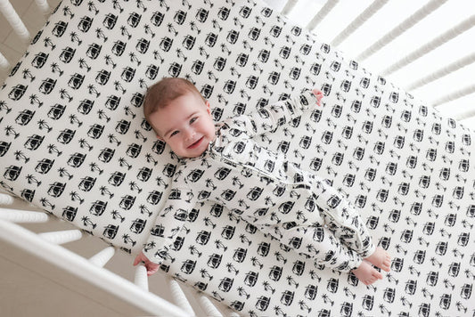 Infant in white romper with black camera and palm tree print, lying in crib.