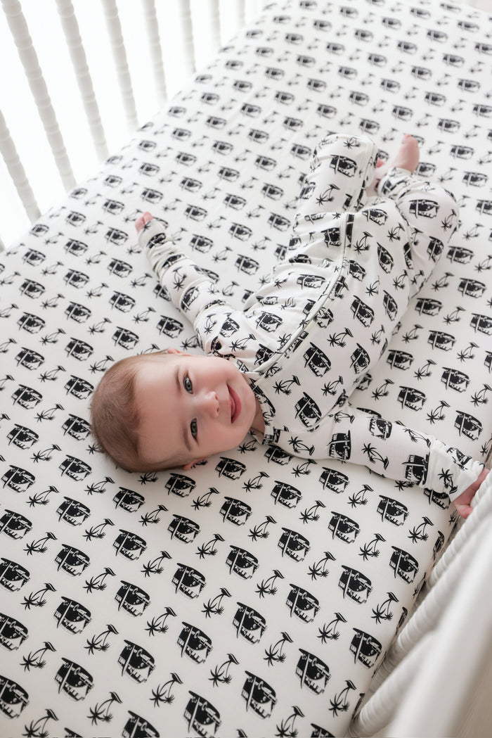 Infant lying on crib sheet wearing white romper with black camper van and palm tree print and pink trim.