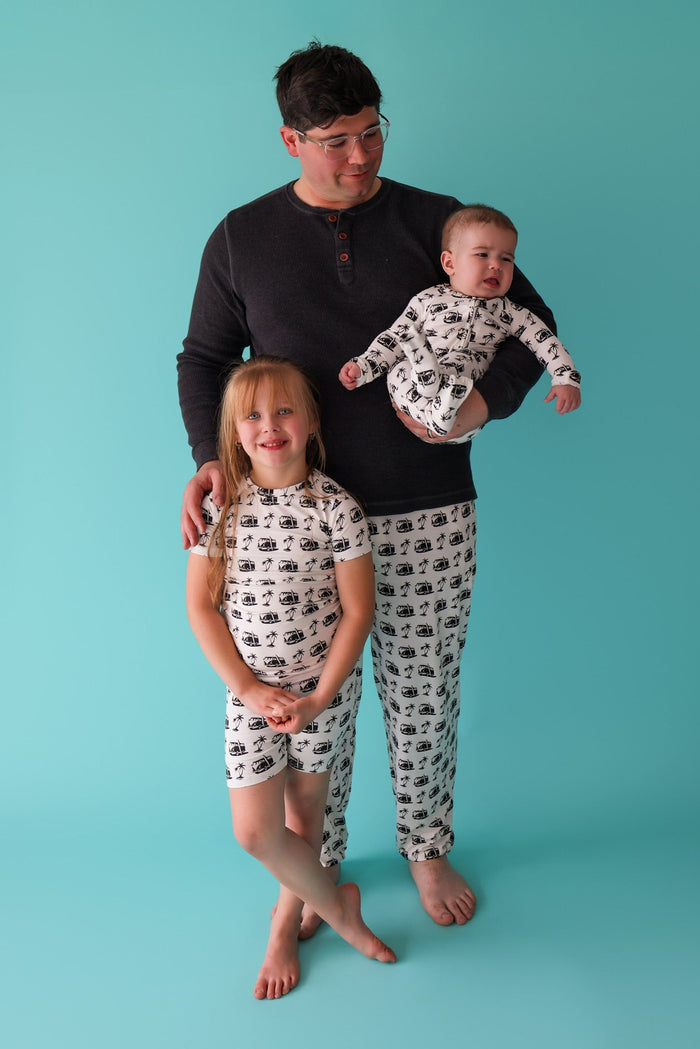 Adult and children wearing matching cream pajamas with black vehicle print pattern against turquoise background.