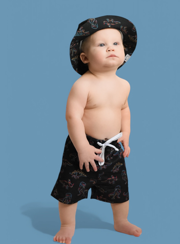 Toddler wearing black swim trunks with colorful dinosaur print and matching swim cap against blue background.