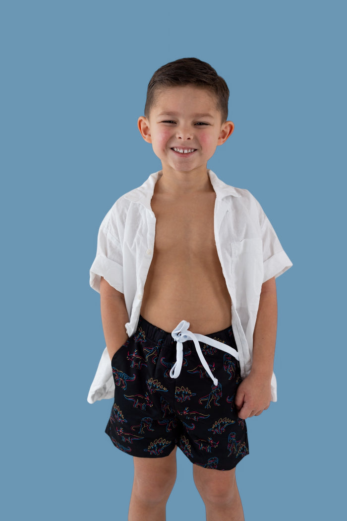Black swim trunks with colorful dinosaur outlines and white drawstring on child model against blue background.
