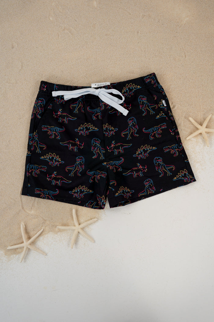 Black swim trunks with multicolored dinosaur outlines and white drawstring, displayed on sand with starfish.