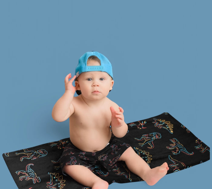 Baby wearing blue towel cap sits on black towel with colorful dinosaur outlines against blue background.