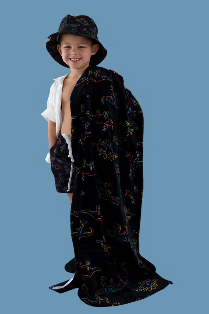 Child wearing black towel with colorful dinosaur outlines, matching bucket hat, draped over white shirt against blue backgrou