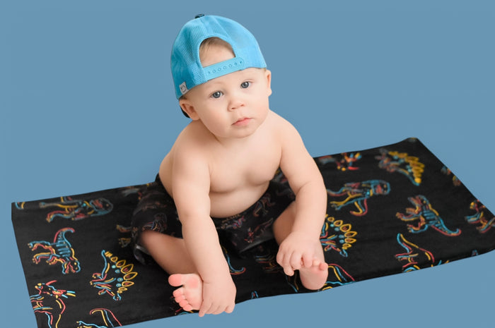Infant wearing light blue towel cap sits on black towel with colorful dinosaur print against blue background.
