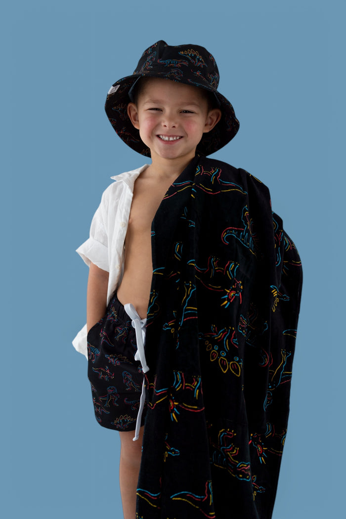 Child wearing black towel with colorful dinosaur outlines and matching bucket hat, draped over white shirt.