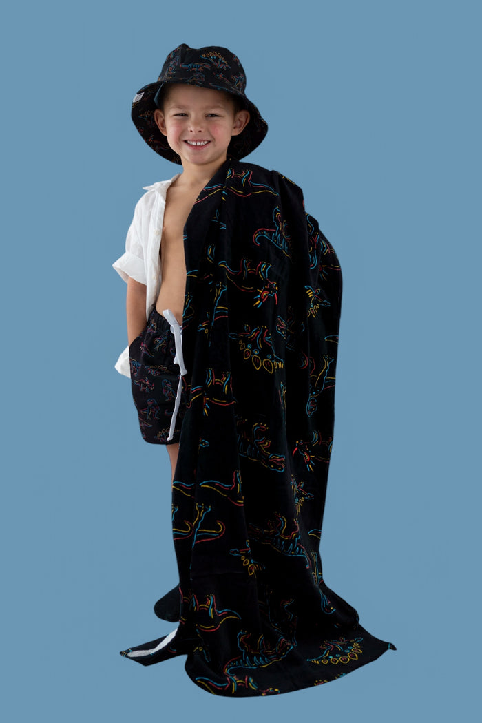 Child wearing black wearable towel with colorful dinosaur outlines, matching bucket hat, against blue background.