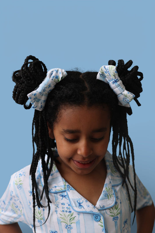 Pair of black hair clips with white fabric bows featuring blue floral print, worn in braided pigtails.