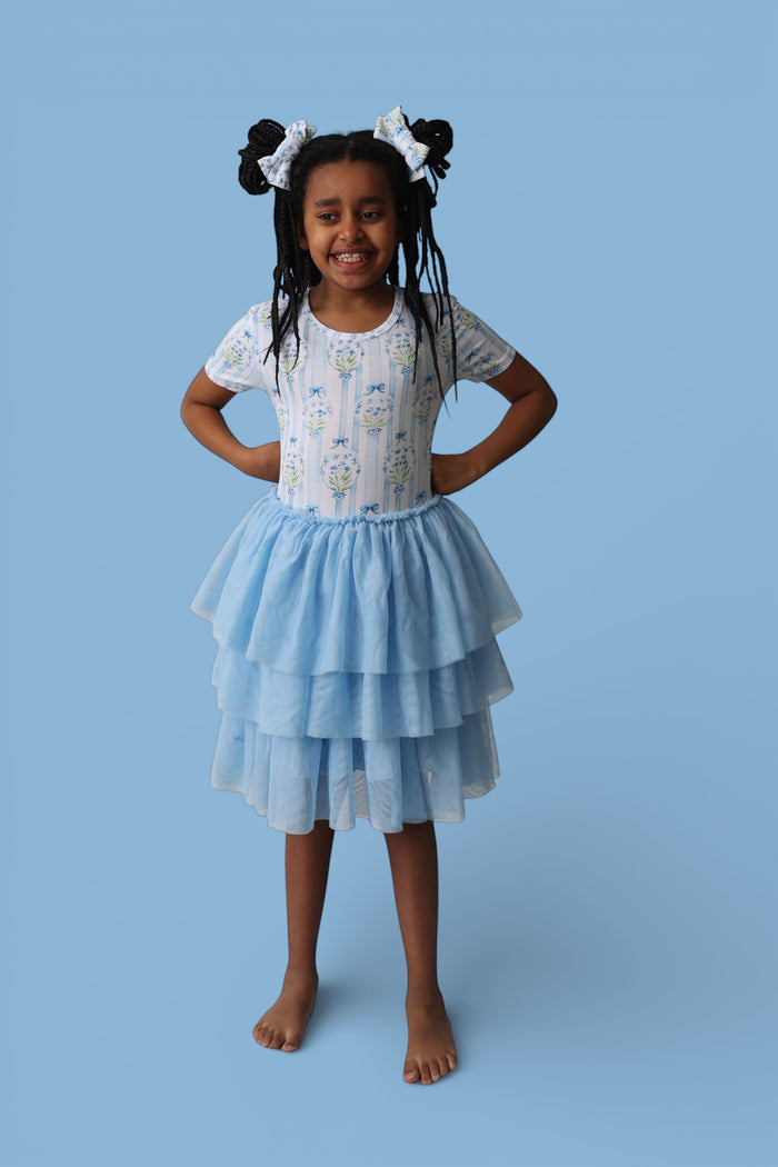 Girl wearing white short-sleeve top with blue floral print and light blue tiered tulle skirt against blue background.