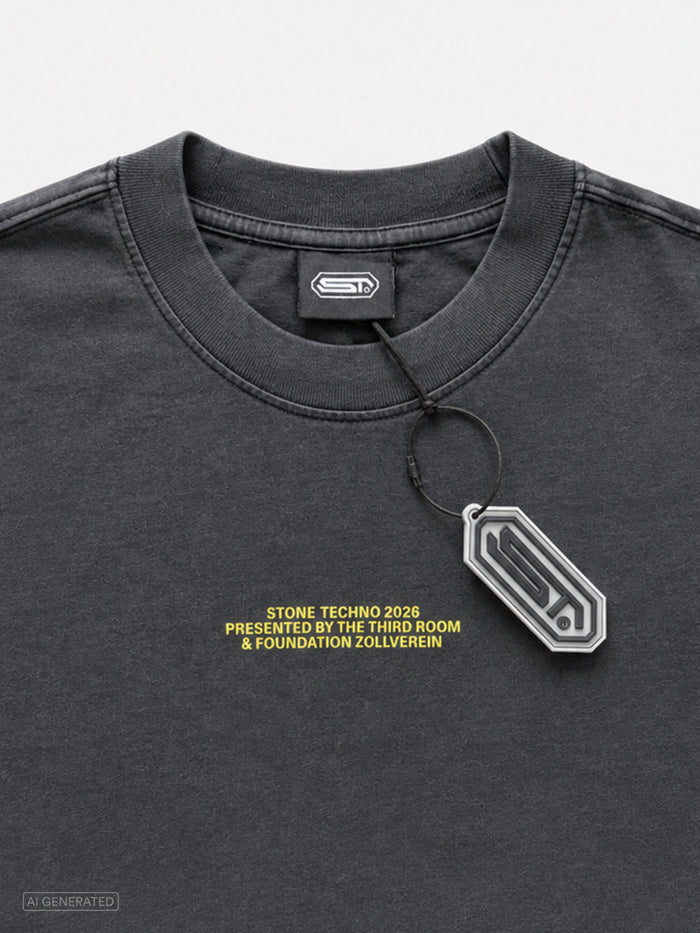 Nahaufnahme des dunkelgrauen T-Shirts mit kleinem gelbem Brustprint „STONE TECHNO 2026 PRESENTED BY THE THIRD ROOM & FOUNDATION ZOLLVEREIN“. Sichtbar sind der gerippte Rundhalskragen, ein schwarzes Nackenlabel mit weißem Logo sowie ein daran befestigter Anhänger. Hintergrund in Hellgrau.