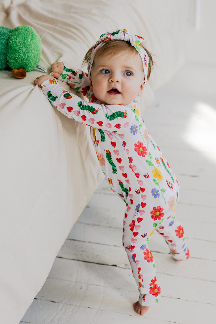 Infant in white romper with colorful floral and heart print, matching headband, lying on white bedding.