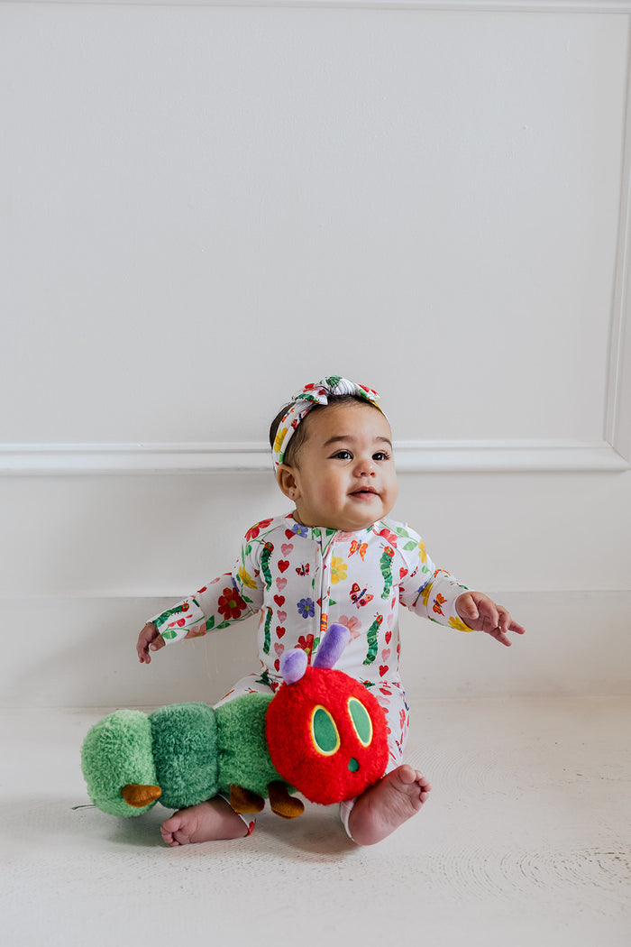 Infant wearing white long-sleeve romper with colorful heart and flower print, matching headband, and green booties.