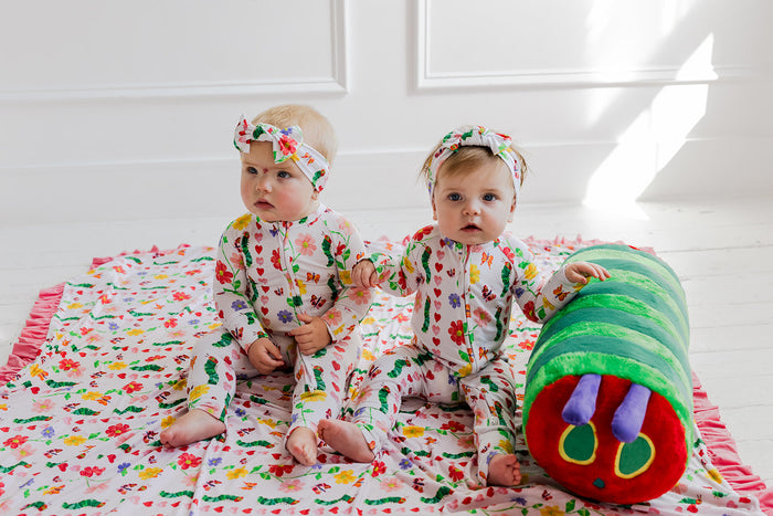 Two babies wearing multicolored floral rompers and matching headbands, sitting on a coordinating patterned blanket.