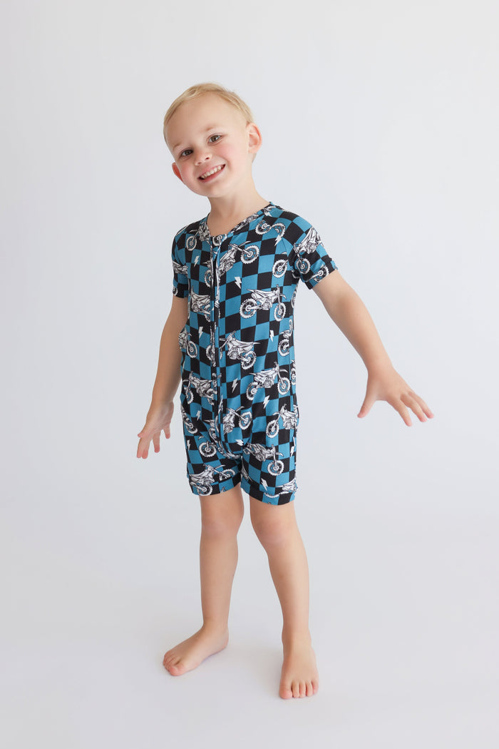 Child wearing short-sleeved romper with blue and black checkered pattern and motorcycle print throughout.