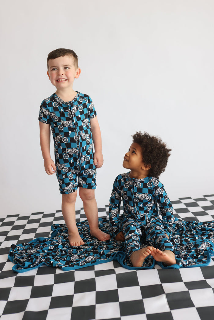 Two children wearing blue and black checkered motorcycle-print pajama sets, one standing and one sitting.