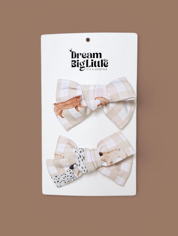 Two gingham bow hair clips with animal designs: dachshund and dalmatian patterns on beige and white checkered fabric.