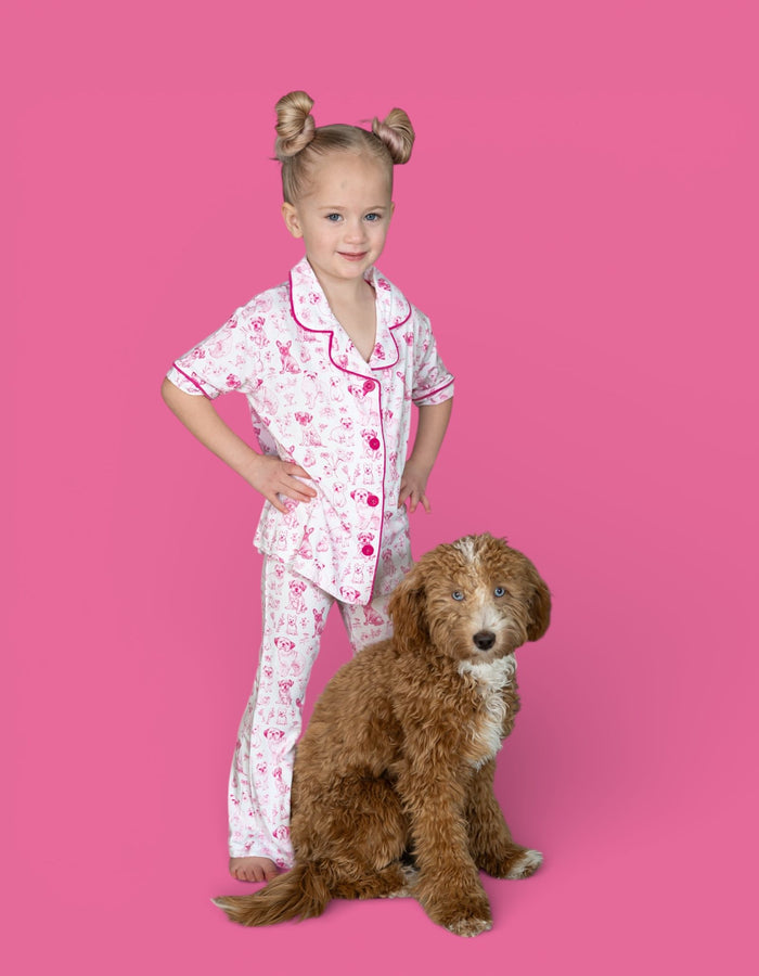 Girl wearing white pajama set with pink puppy toile print and matching flare pants, standing next to brown dog.