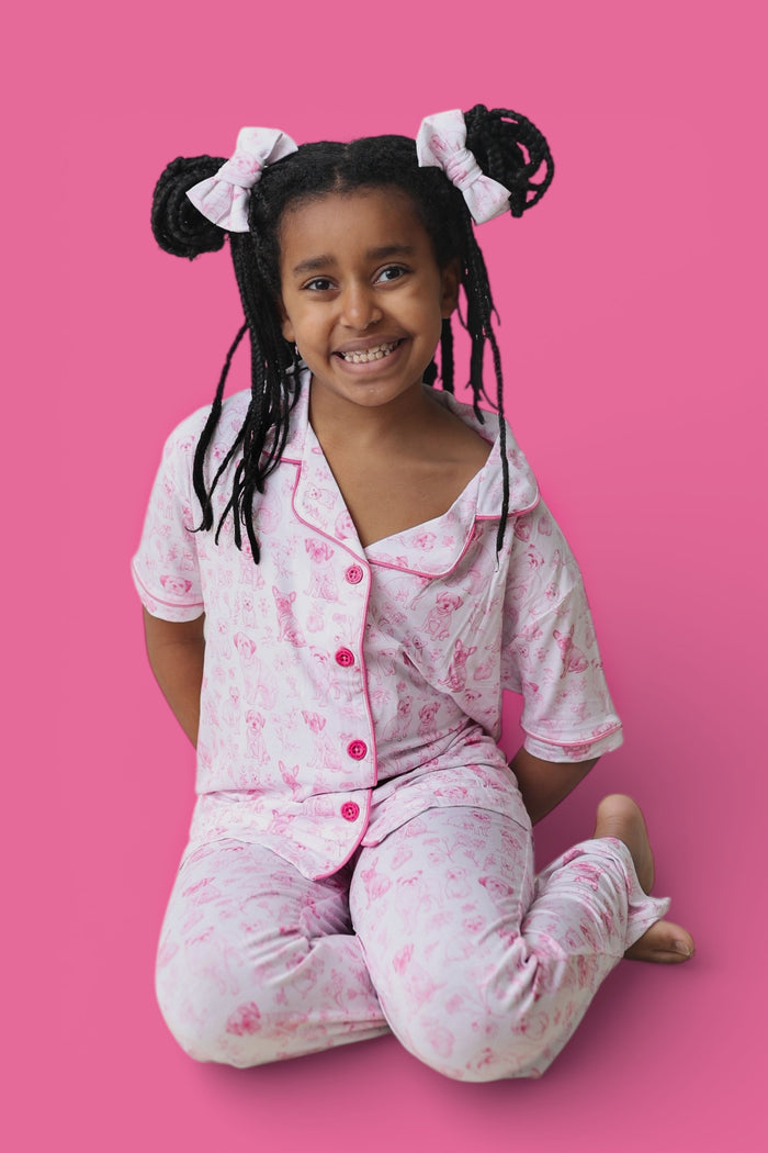 Girl wearing pink toile puppy print short-sleeve pajama set with red buttons and flared pants legs.