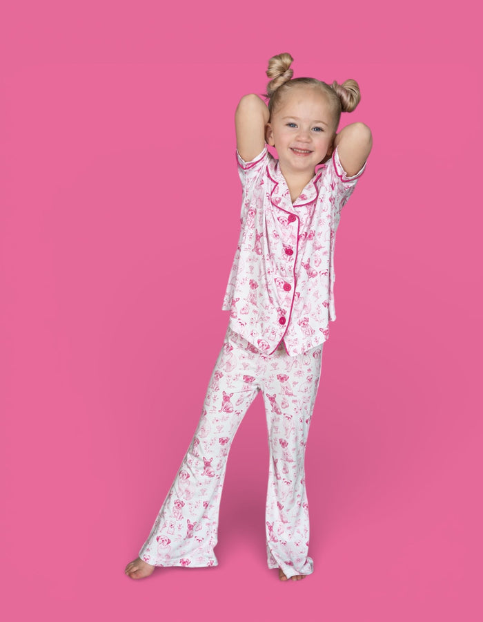 Girl wearing pink and white puppy toile print pajama set with flared pants and matching shirt against pink background.